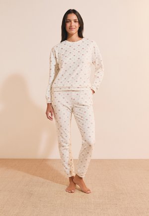 Donna in piedi a piedi nudi con top a maniche lunghe floreale color crema e pantaloni da jogging abbinati su sfondo neutro.