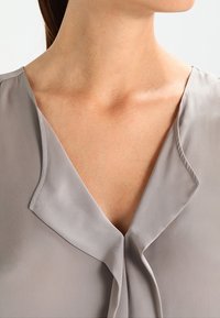 Blusa grigia e setosa con scollo a V e morbido dettaglio a piega sul colletto. Il tessuto ha una leggera lucentezza e una texture liscia.