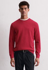 Roter Strickpullover mit einem strukturierten Muster, runder Ausschnitt und kontrastierendem weißen Saum. Getragen über dunklen, maßgeschneiderten Hosen.