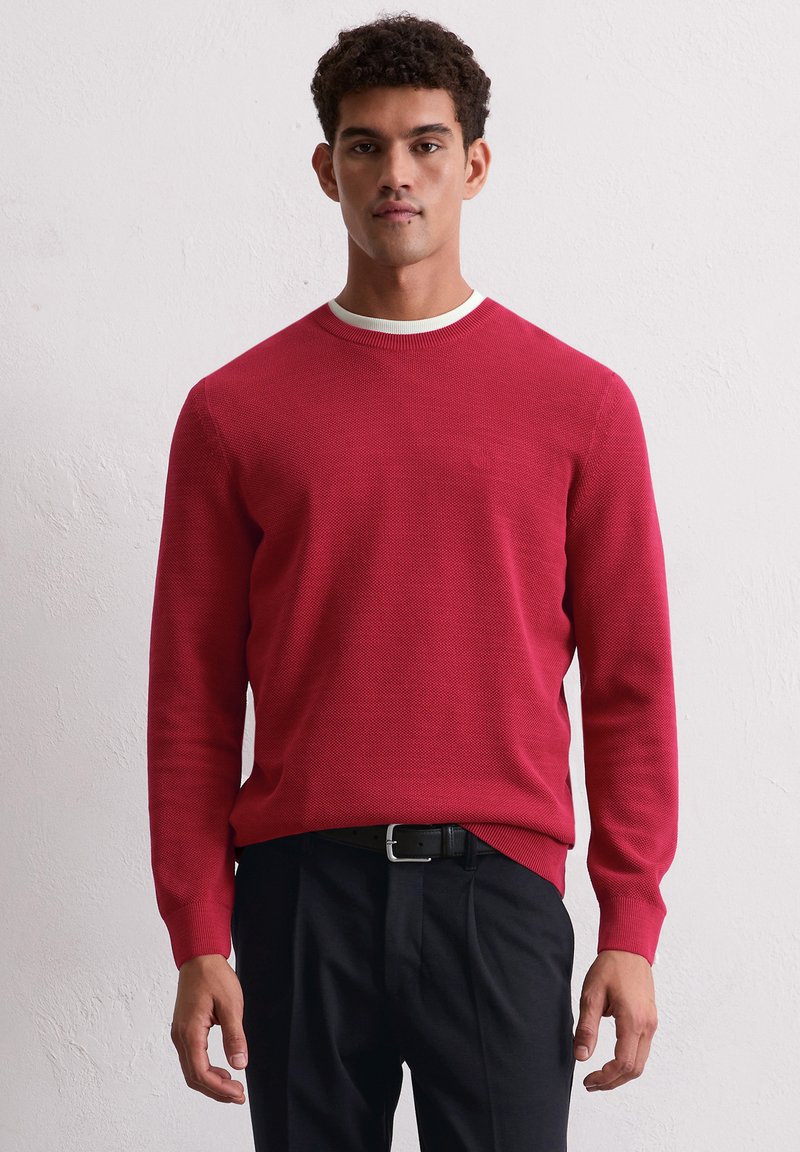Roter Strickpullover mit einem strukturierten Muster, runder Ausschnitt und kontrastierendem weißen Saum. Getragen über dunklen, maßgeschneiderten Hosen.