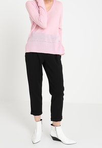 Maglione rosa chiaro con scollatura a V e texture a coste, abbinato a pantaloni neri arrotolati e stivaletti bianchi a punta con cerniere.