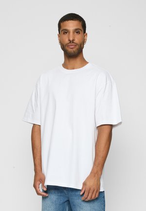 Muž s krátkymi vlasmi a náušnicami, nosiaci jednoduché biele voľné tričko a modré džínsy, stojaci na pozadí s jednoliatou sivou farbou.