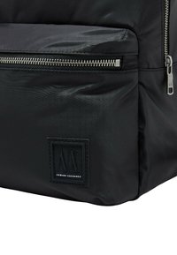 Mochila negra de nailon con una textura suave, con herrajes de cremallera plateados y una chapa de logo en relieve. Diseño estructurado con un bolsillo frontal.