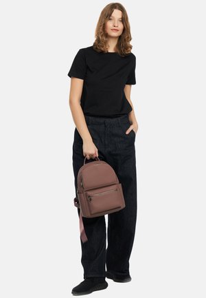Sac à dos brun texturé avec deux fermetures éclair avant, dessus arrondi et bretelles réglables, associé à un t-shirt noir et un pantalon large et foncé.