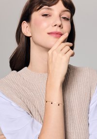 Złota bransoletka łańcuszkowa z trzema małymi czarnymi serduszkami noszona na nadgarstku modelki. Modelka ma włosy brązowe do ramion i nosi beżowy sweter.