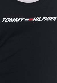 Musta puuvillapaita, jossa on valkoinen "TOMMY" ja "HILFIGER" -logo sekä punainen ja sininen suorakaidekorostus, pyöreä pääntie ja sileä pinta.