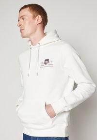Sweat à capuche blanc en tissu doux, avec une poche frontale, des cordons de serrage et un logo brodé. Le modèle porte un jean bleu.