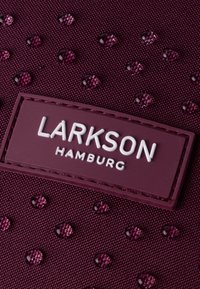 Burgundi színű, texturált felületű szövet, amelyen fehérrel gravírozott "LARKSON HAMBURG" címke és fényes lila szegecsek találhatók.
