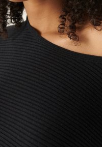 Mèches de cheveux bouclés reposant sur l'épaule, portant un haut noir côtelé à épaules dénudées, vue rapprochée du décolleté et de la texture du tissu.