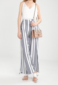 Top bianco senza maniche con scollo a V, abbinato a pantaloni larghi a righe blu navy e bianche a vita alta. Tiene in mano una piccola borsa rosa a trama.