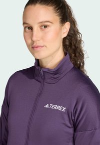Giacca zip-up viola con colletto alto, realizzata in tessuto testurizzato e con il logo bianco "TERREX" sul petto sinistro. Nessun motivo o decorazione visibile.