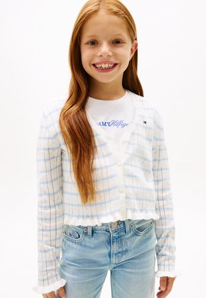 Lachend roodharig meisje met een wit-blauw gestreepte cardigan over een wit Tommy Hilfiger-shirt en lichtblauwe spijkerbroek.