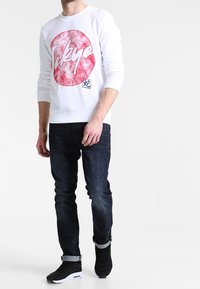 Sweatshirt blanc avec un graphique circulaire rouge portant le texte "Tokyo" en police audacieuse, associé à un jean bleu foncé et des baskets noires.