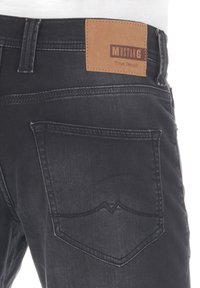 Pantalones vaqueros de mezclilla negra con una etiqueta de cuero que dice "Mustang True Denim". Presenta un distintivo cosido en los bolsillos traseros y una textura de tela suave.
