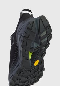 Mustan urheilukengän pohja, jossa on syvät urat, tekstuuripinta ja merkittävä keltainen yksityiskohta kannan lähellä. Kevyt muotoilu.