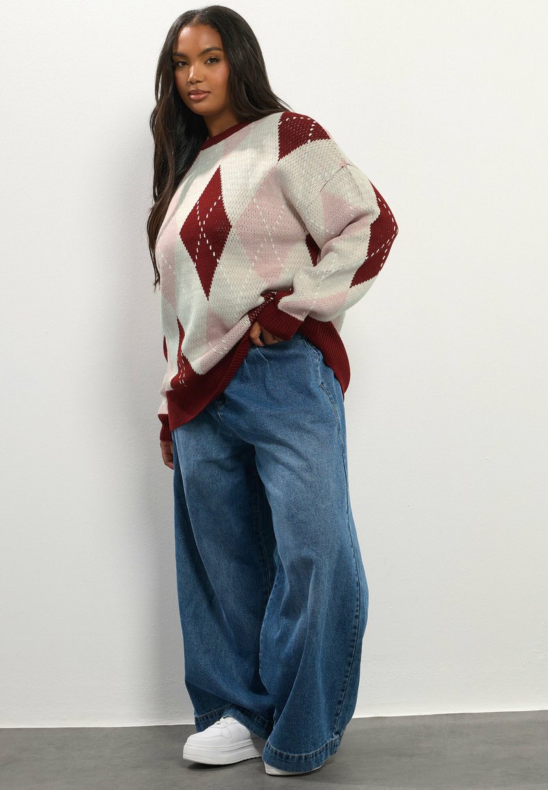 Neule, jossa on timanttikuvio punaisena, vaaleanpinkkinä ja kermanvärisenä, yhdistettynä leveälahkeisiin sinisiin farkkuihin ja valkoisiin tennareihin neutraalilla taustalla.