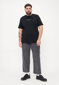 Zwarte katoen t-shirt met korte mouwen, voorzien van witte tekst op de voorkant. Gecombineerd met grijze ribfluwelen broek en zwarte veterschoenen.