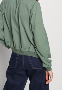 Zelená, lehká mikina se staženým lemem a volným střihem, ve spojení s tmavě modrými džíny s bílým prošitím na kapsách.