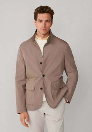 Chaqueta marrón con cuello y tres botones, que cuenta con dos bolsillos delanteros. Tejido ligeramente texturizado, usada sobre un suéter blanco y una camisa amarilla.