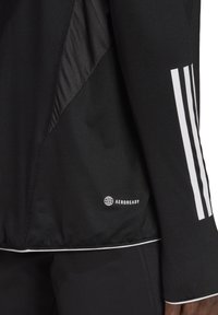 Giacca sportiva nera con pannelli in rete, dettagli bianchi e un logo. Il design presenta tre strisce bianche lungo la manica.