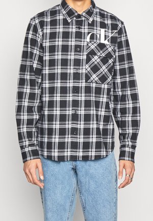 Homme portant une chemise à manches longues à carreaux noirs et blancs avec une poche avant et un jean bleu délavé, debout devant un fond uni.