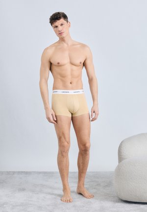 Uomo in piedi a piedi nudi su un tappeto che indossa slip boxer beige Lacoste con fascia elastica bianca, accanto a una poltrona imbottita curva bianca.