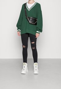 Pull vert tricoté oversize avec des accents blancs, jean skinny noir déchiré et baskets blanches épaisses. Sac bandoulière noir avec chaîne.