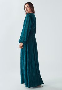 Tyrkysové dlouhé šaty se zmačkanou texturou, dlouhými nabíranými rukávy a volným střihem, zobrazené z bočního úhlu. Design až na podlahu bez viditelného kování.