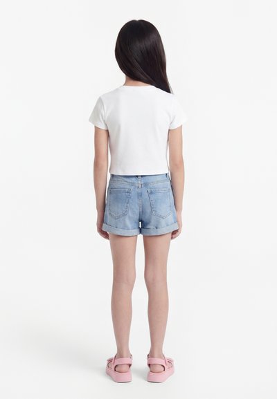 Jeune fille debout, dos à la caméra, portant un t-shirt blanc à manches courtes, un short en jean retroussé et des sandales roses sur fond blanc.