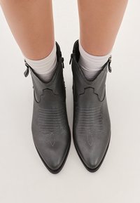 Botas de cuero gris hasta el tobillo con punteras afiladas, diseño con bordes ondulados, costuras en contraste y cremalleras laterales; combinadas con calcetines blancos.