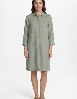Femme debout portant une robe chemise vert clair à manches longues, longueur genoux, avec un col et des boutons cachés sur le devant, sur fond blanc.