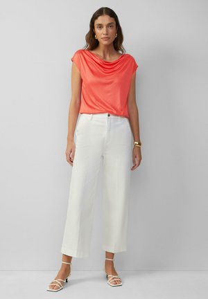 Korallenfarbener Rollkragen-Top, leichtes Gewebe. Weiße, weit geschnittene Hosen, taillierte Passform, glatte Textur. Weiße Sandalen mit Riemchen und kleinem Absatz.