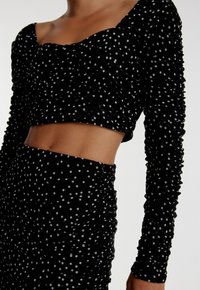 Zwarte cropped top met lange mouwen, voorzien van zilveren stippen en gerimpelde textuur. Coördineert met een bijpassende gerimpelde rok.