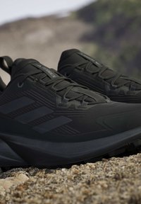 Zwarte Adidas Terrex trailrunning schoenen rustend op rotsachtige grond met een wazige buitenachtergrond.