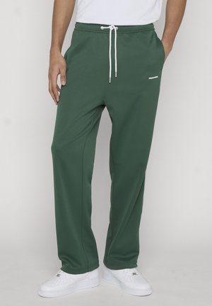 Uomo che indossa pantaloni verdi scuro a taglio ampio con coulisse bianca, sneakers bianche e una camicia bianca infilata, in piedi davanti a uno sfondo neutro.