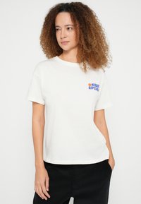Hvid bomuld t-shirt med korte ærmer og rund hals, med en farverig logo på venstre bryst. Modellen bærer mørke bukser. Baggrunden er ensfarvet.