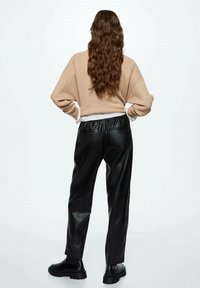 Beżowy sweter z dzianiny, czarne spodnie z ekoskóry i czarne botki za kostkę. Długie, falujące włosy opadają na plecy. Minimalistyczne tło.