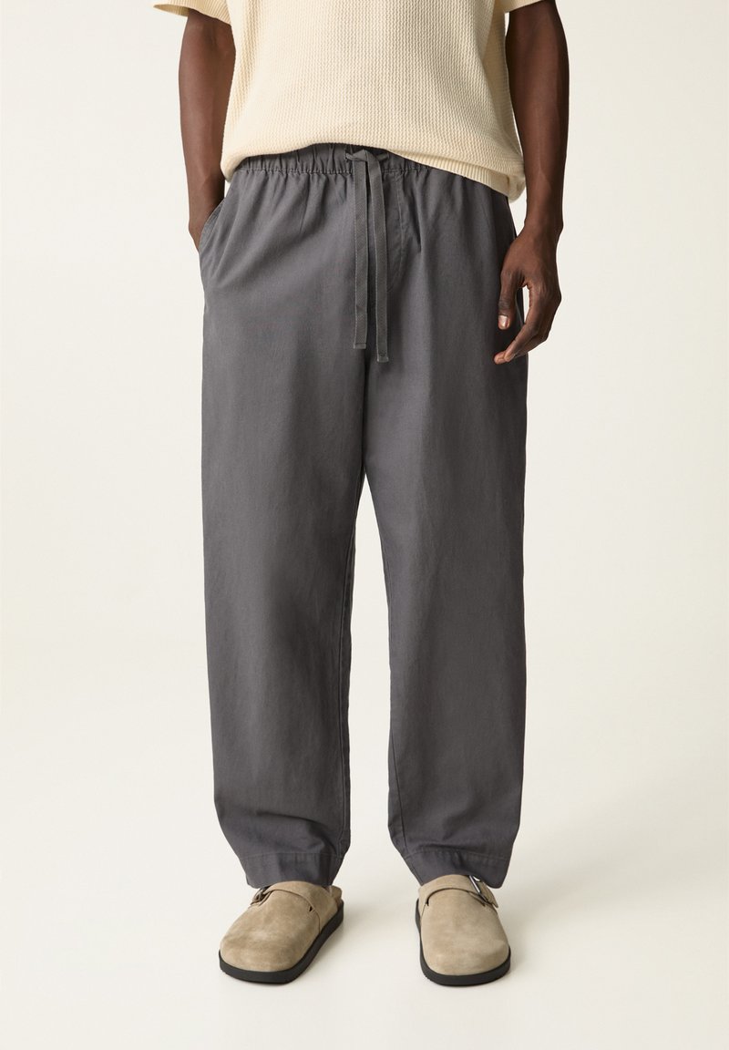 Graue Leinenhosen mit einem elastischen Bund und Kordelzug. Lockere Passform, Seitentaschen und schmal zulaufende Knöchel, kombiniert mit beigen Slippern.