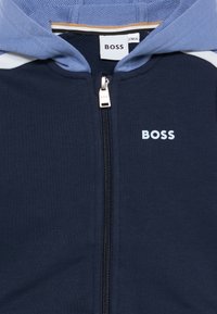 Φούτερ με φερμουάρ σε ναυτικό μπλε με κουκούλα σε ανοιχτό γαλάζιο, διαθέτει ασημί φερμουάρ, ριγέ λευκά διακοσμητικά και το λογότυπο "BOSS" τυπωμένο λευκό. Ομαλή υφή.