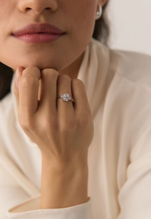 Close-up of a woman’s hand resting on her chin, wearing a silver ring with a large square-cut clear gemstone.