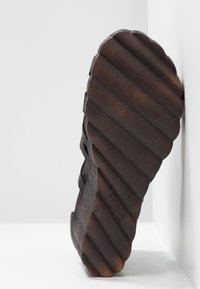 Semelle d'une chaussure marron foncé avec des rainures diagonales profondes, présentée à la verticale sur un fond blanc, mettant en valeur la texture et le motif de la bande de roulement.