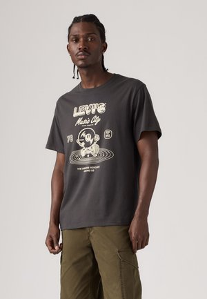 Homme aux cheveux tressés portant un t-shirt graphique sombre Levi's Music City et un pantalon cargo olive, posant devant un fond clair uni.