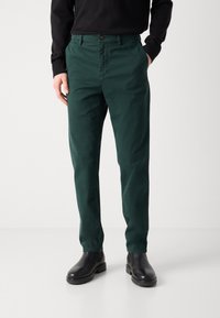 Chinos de algodón verde con un corte recto, que cuentan con bolsillos laterales y cierre de botón. Llevados con botas de cuero negras para un contraste.