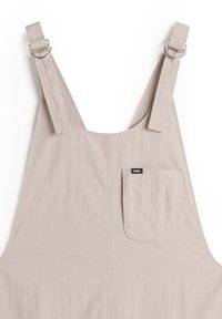 Beige Latzhose aus weichem Stoff mit verstellbaren Trägern mit Metallringen, einer aufgesetzten Tasche und einer dezenten strukturierten Oberfläche.
