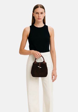 Borsa in suede marrone con forma arrotondata, manico in pelle e dettagli in metallo argento. Il modello indossa un top senza maniche nero e pantaloni bianchi.