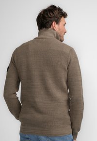 Suéter marrón de punto con un cuello alto, tejido texturizado y dobladillo acanalado. Presenta un respaldo liso y un parche con el logo en la manga izquierda.