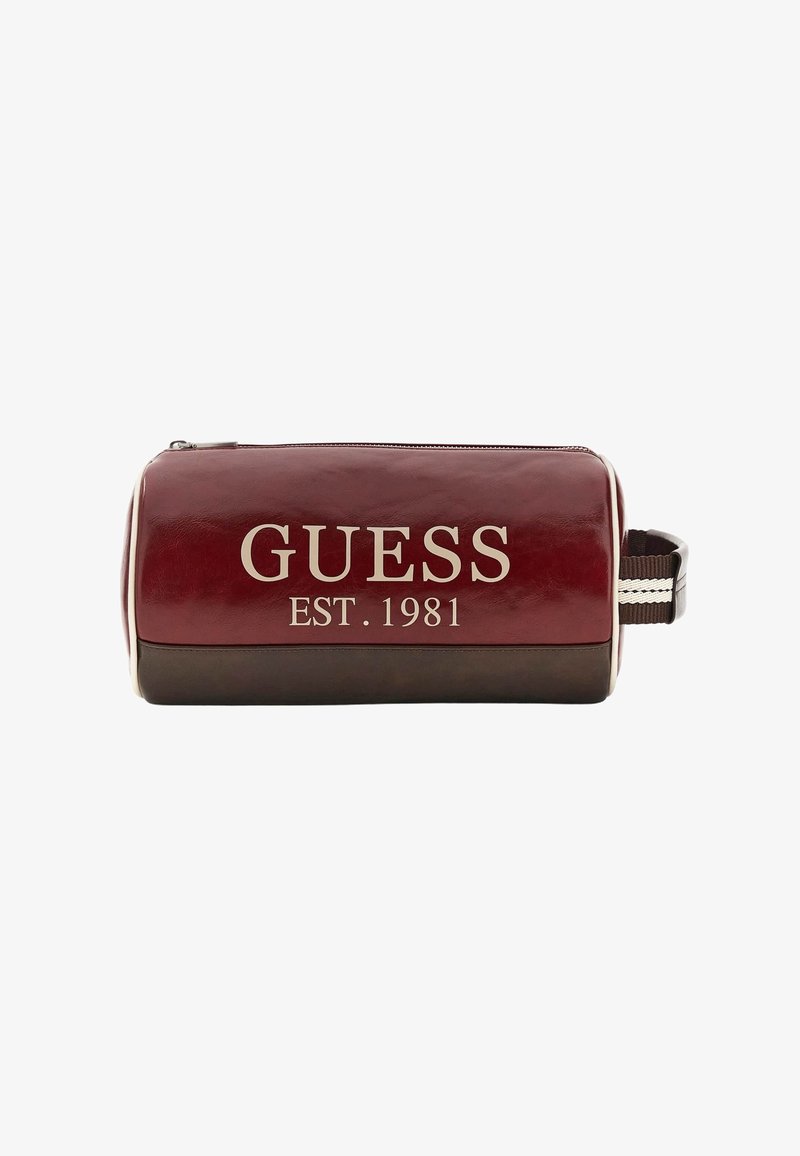 Trousse de toilette rectangulaire en cuir marron rouge et brun avec fermeture éclair, poignée latérale et inscription "GUESS EST. 1981" imprimée en blanc sur le devant.
