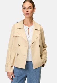 Beige dubbelknäppt jacka med slagkrage, stora knappar, sidofickor och långa ärmar, kombinerad med en vit topp och denimpants.