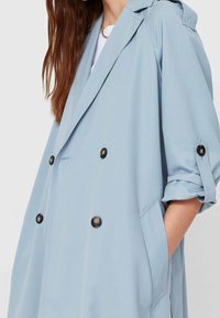 Jasnoniebieska dwurzędowa marynarka z czarnymi guzikami, szerokim kołnierzem i podwiniętymi rękawami ozdobionymi guzikami. Miękki materiał i luźny krój.