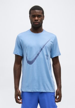 Uomo che indossa una T-shirt Nike azzurro chiaro con grande logo swoosh sbiadito e pantaloncini blu reale, in piedi contro uno sfondo bianco semplice.